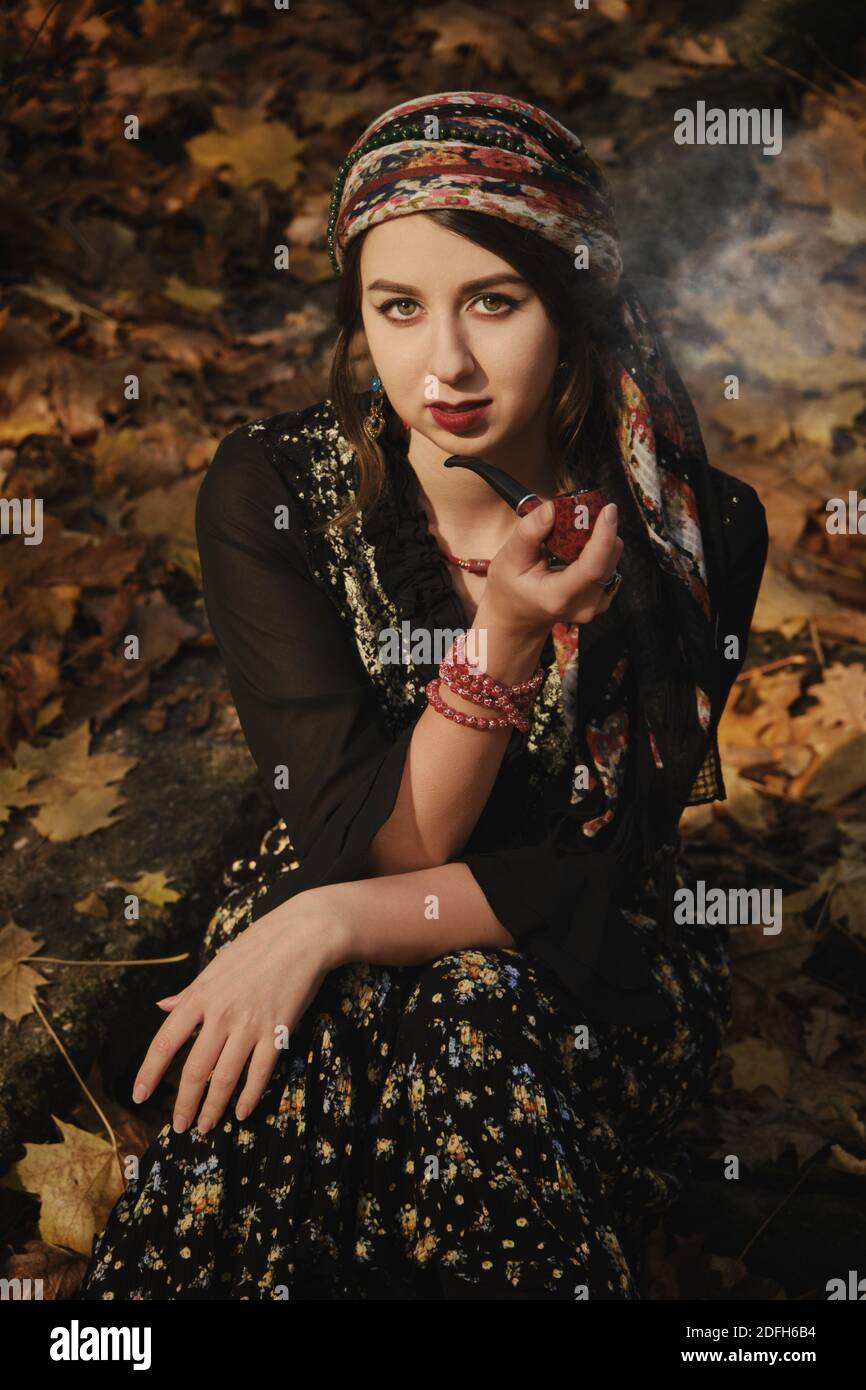 young gypsy woman smoking pipe in autumn forest, looking at camera ...