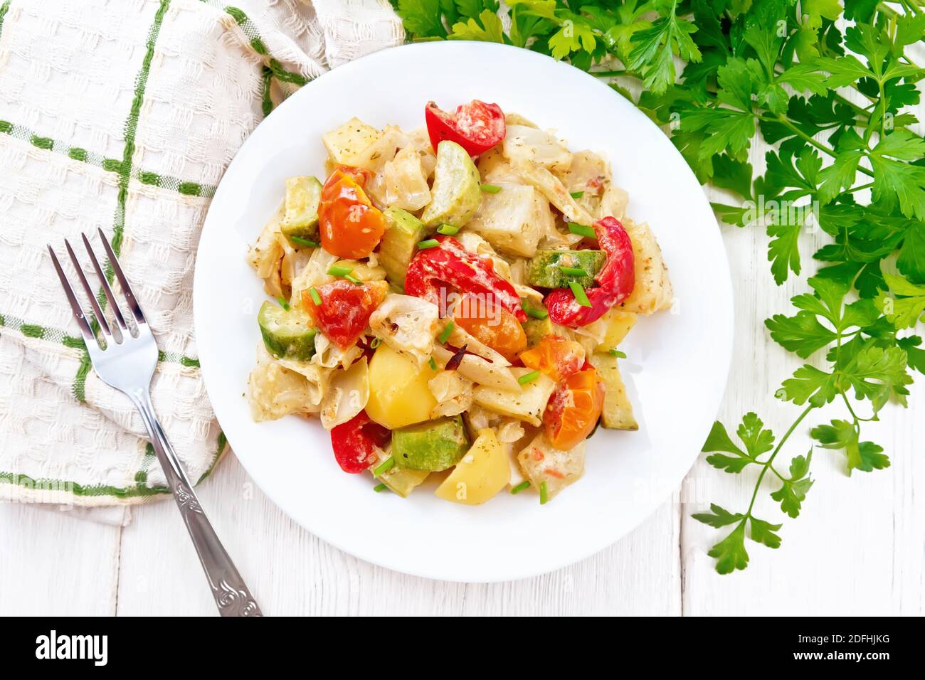 Vegetable ragout with zucchini, cabbage, potatoes, tomatoes and bell peppers in creamy sauce in plate, napkin, parsley and a fork on the background of Stock Photo