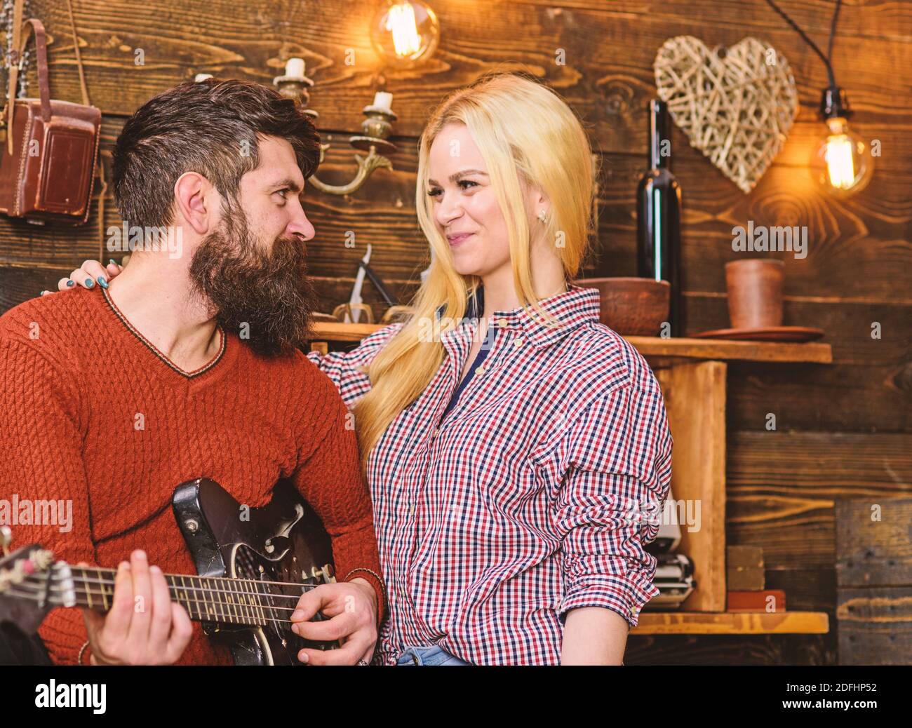 Romantic evening concept. Couple in love spend romantic evening in warm atmosphere. Couple in wooden vintage interior enjoy guitar music. Lady and man with beard on happy faces hugs and plays guitar. Stock Photo