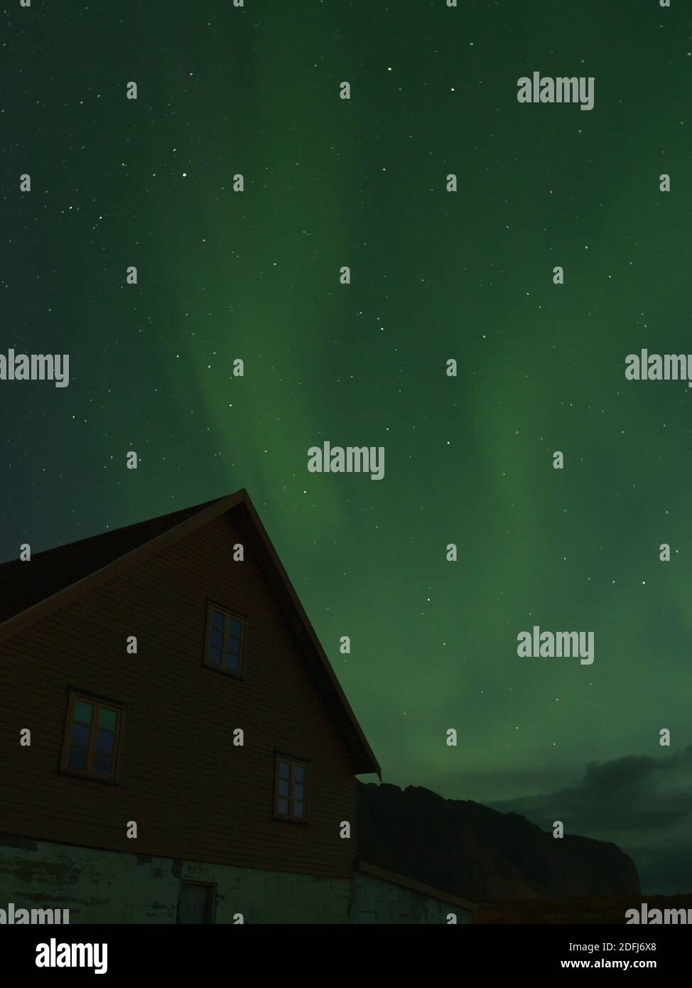 Stunning upright view of night sky with bright green shimmering polar lights (aurora borealis) above old house on Vestvågøy island, Lofoten, Norway. Stock Photo