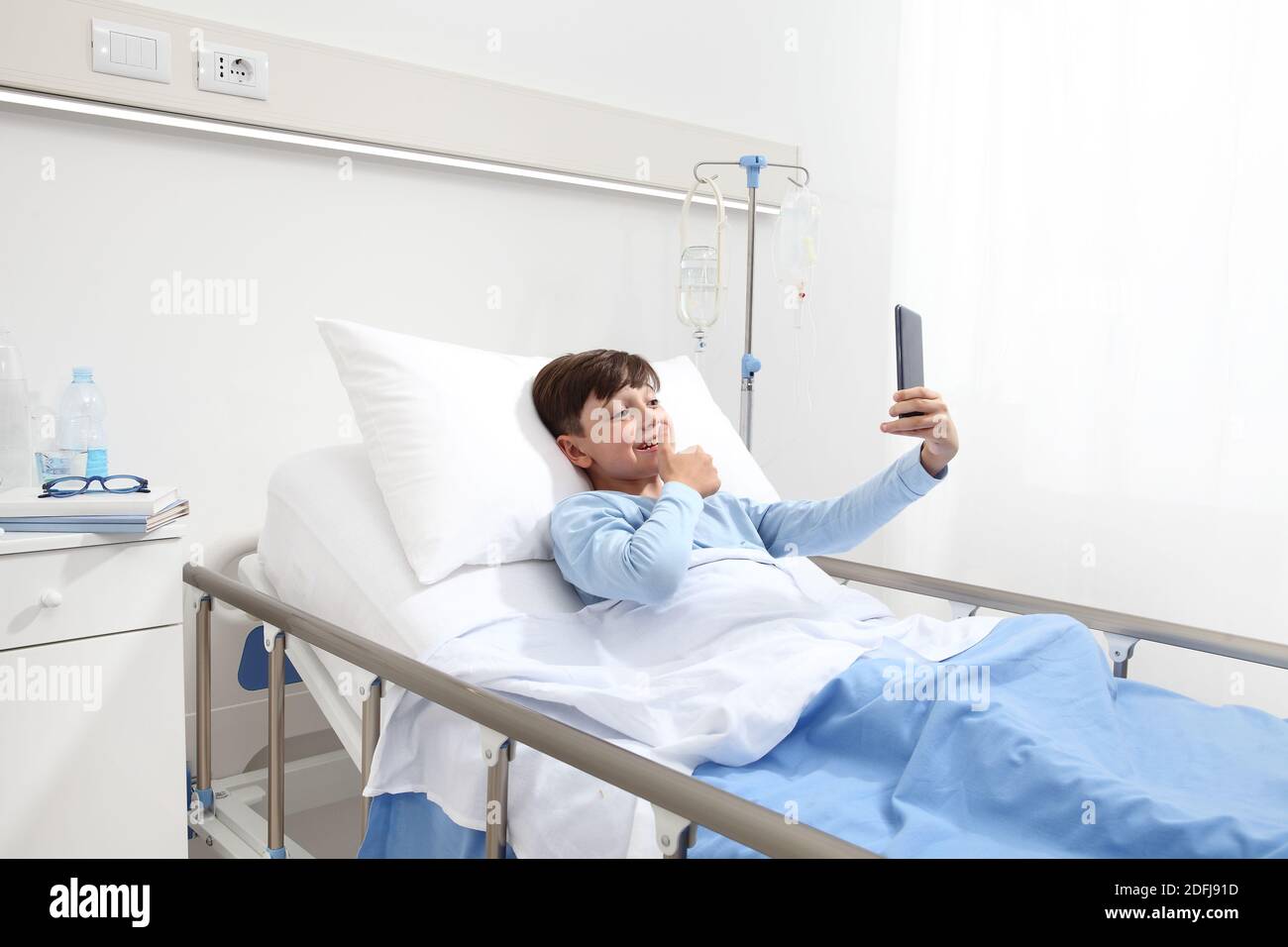 Happy child lying in bed alone in hospital room taking selfie with