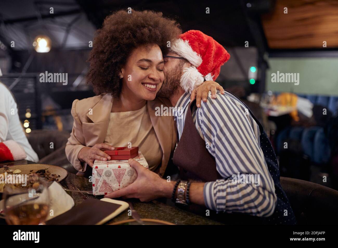 beardy caucasian guy kissing in cheek afro-american female, whispering to ear , giving her a christmas present, wearing santa hat Stock Photo