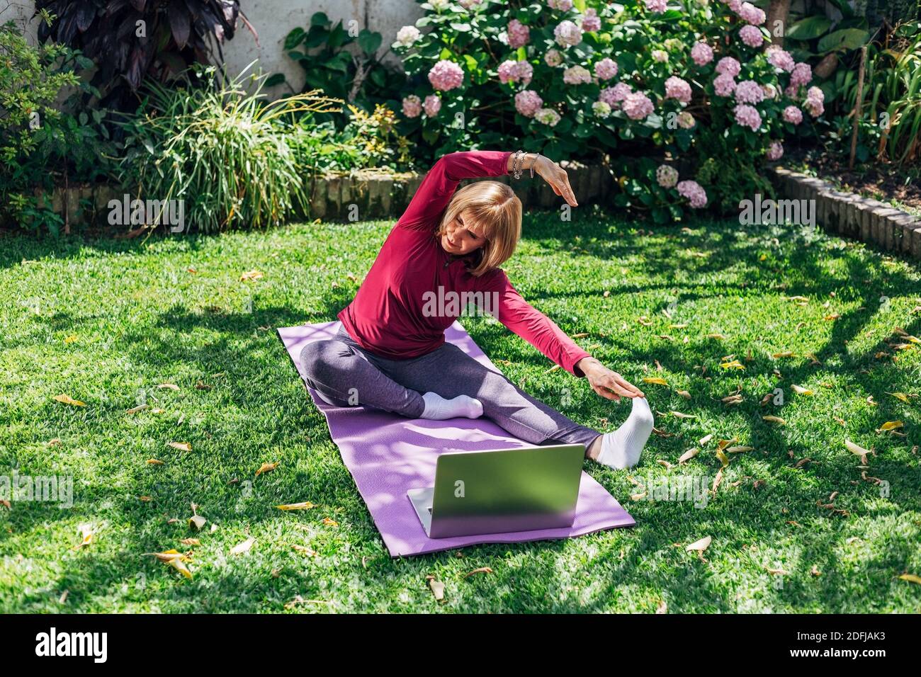Online sport fitness yoga training workout, senior woman in the garden of her home doing exercises on pilates mat opposite laptop at home. Stock Photo
