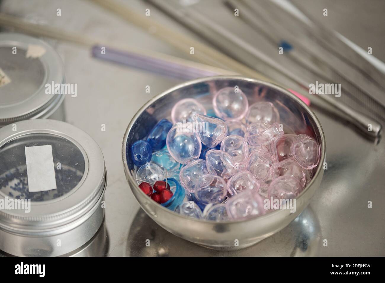 Metallic bowl full of glass workpieces of various shapes and colors standing on workplace of professional lampworker among other supplies Stock Photo