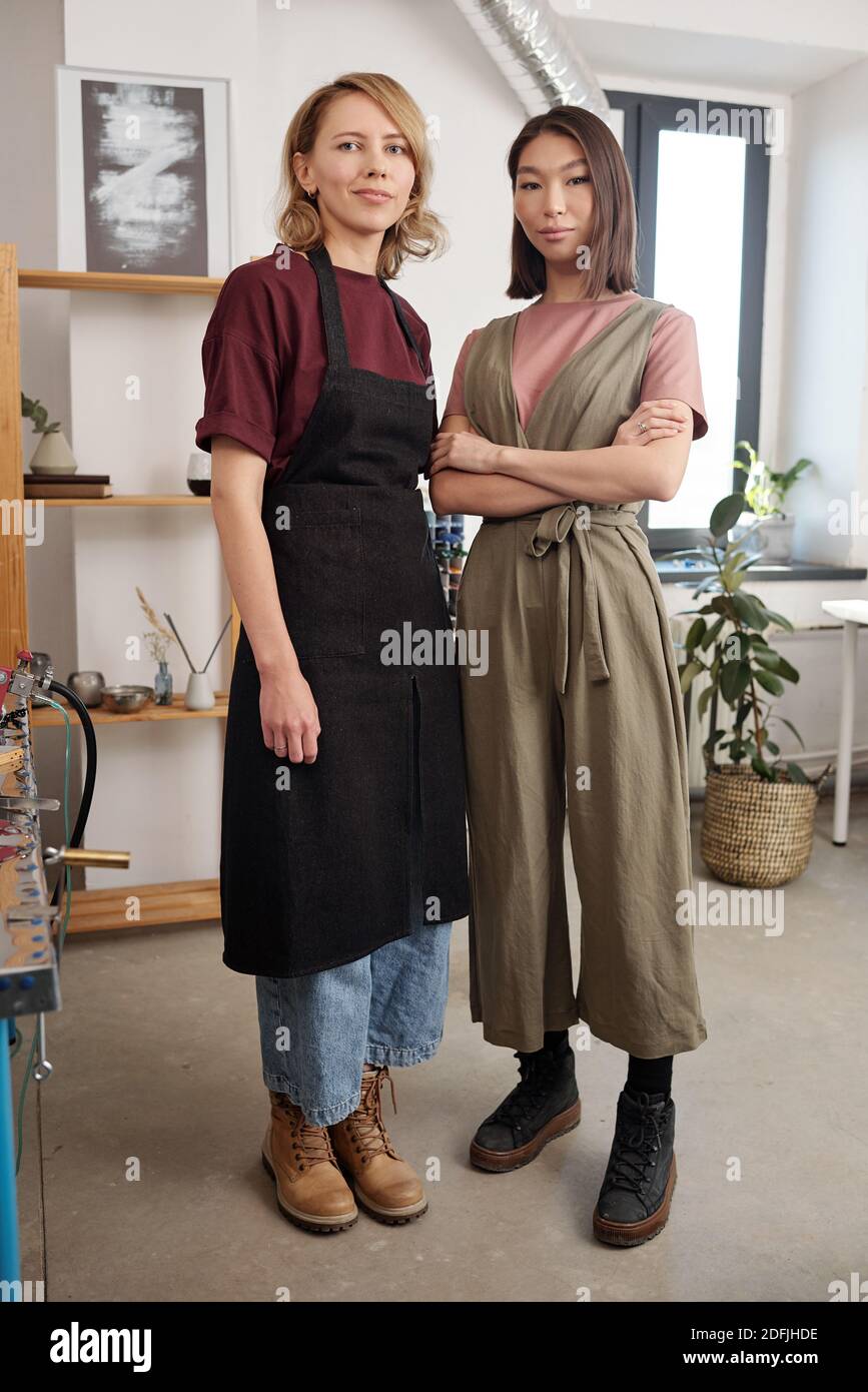 Two young intercultural female artisans in workwear standing next to one another in front of camera in large workshop and looking at you Stock Photo