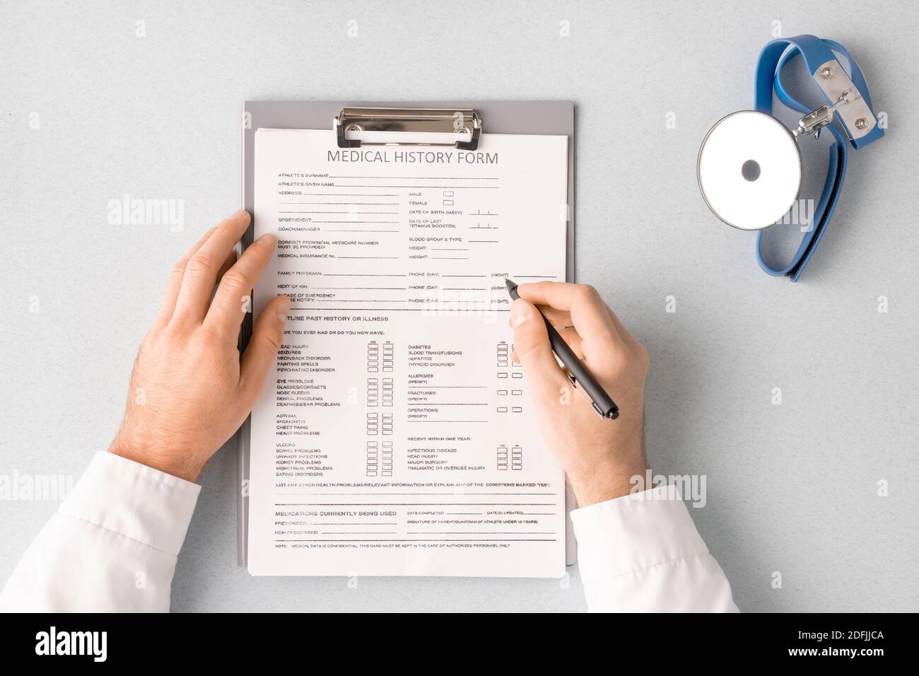 Overview of doctor hands with pen over medical history form in clipboard filling in personal information of patient with clinic tool near by Stock Photo