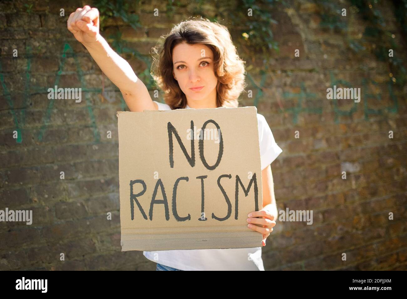 Upset young protesting woman in white shirt holds protest sign broadsheet placard with slogan 'No racism' for public demonstration on stone wall Stock Photo