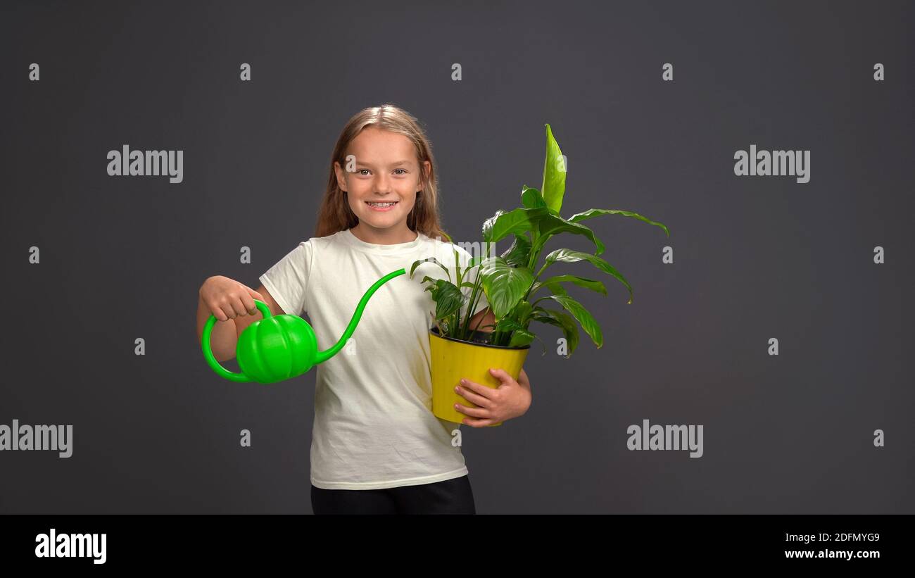 Little girl watering a flower in pot using a funny watering can while holding everything in her hands. Girl wearing white t shirt smiling at camera Stock Photo