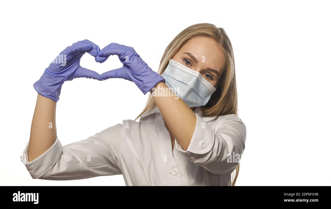 Beautiful young nurse in white robe and medical mask shows love sign with her hands lifting up, looking at the camera. Isolated on white background Stock Photo