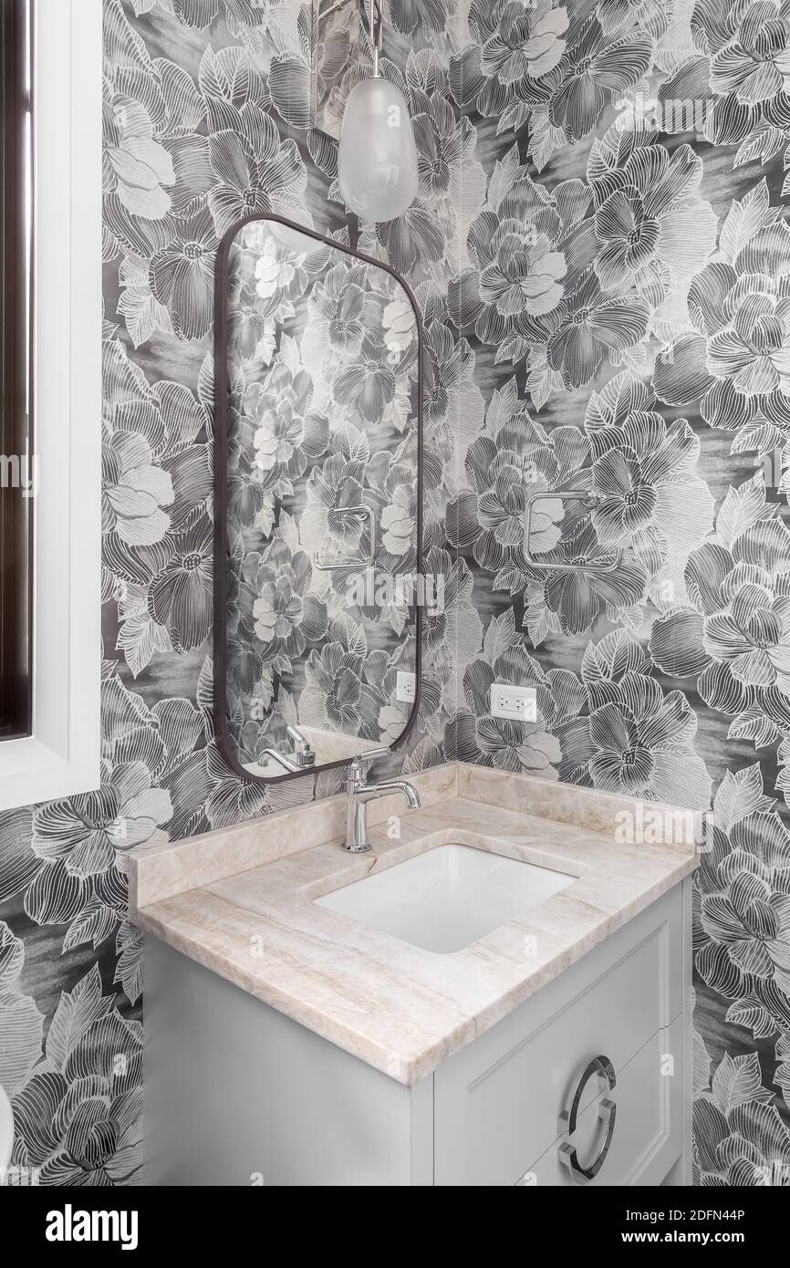 A modern bathroom with a patterned wallpaper, grey vanity and marble counter top, and a beautiful light fixture above the bronze mirror. Stock Photo