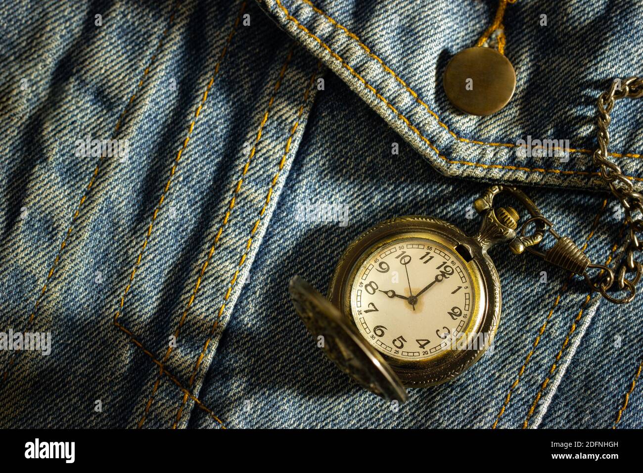Vintage pocket watch is placed over an old blue denim shirt and the morning sun shines down in the top right corner. The concept of time importance. C Stock Photo