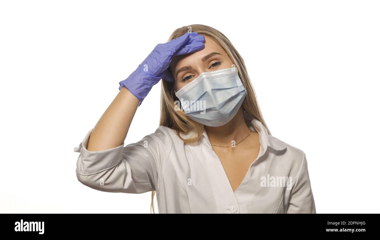 Young doctor in a medical mask is experiencing a headache at work, massaging the lobe of the head, looking at the camera. Isolated on white background Stock Photo
