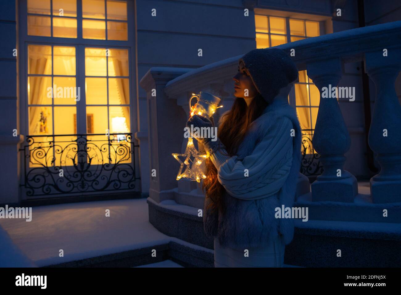 Christmas Eve portrait. Woman in front of her house in Christmas night, surrounded by snowflakes and warm lights. Stock Photo