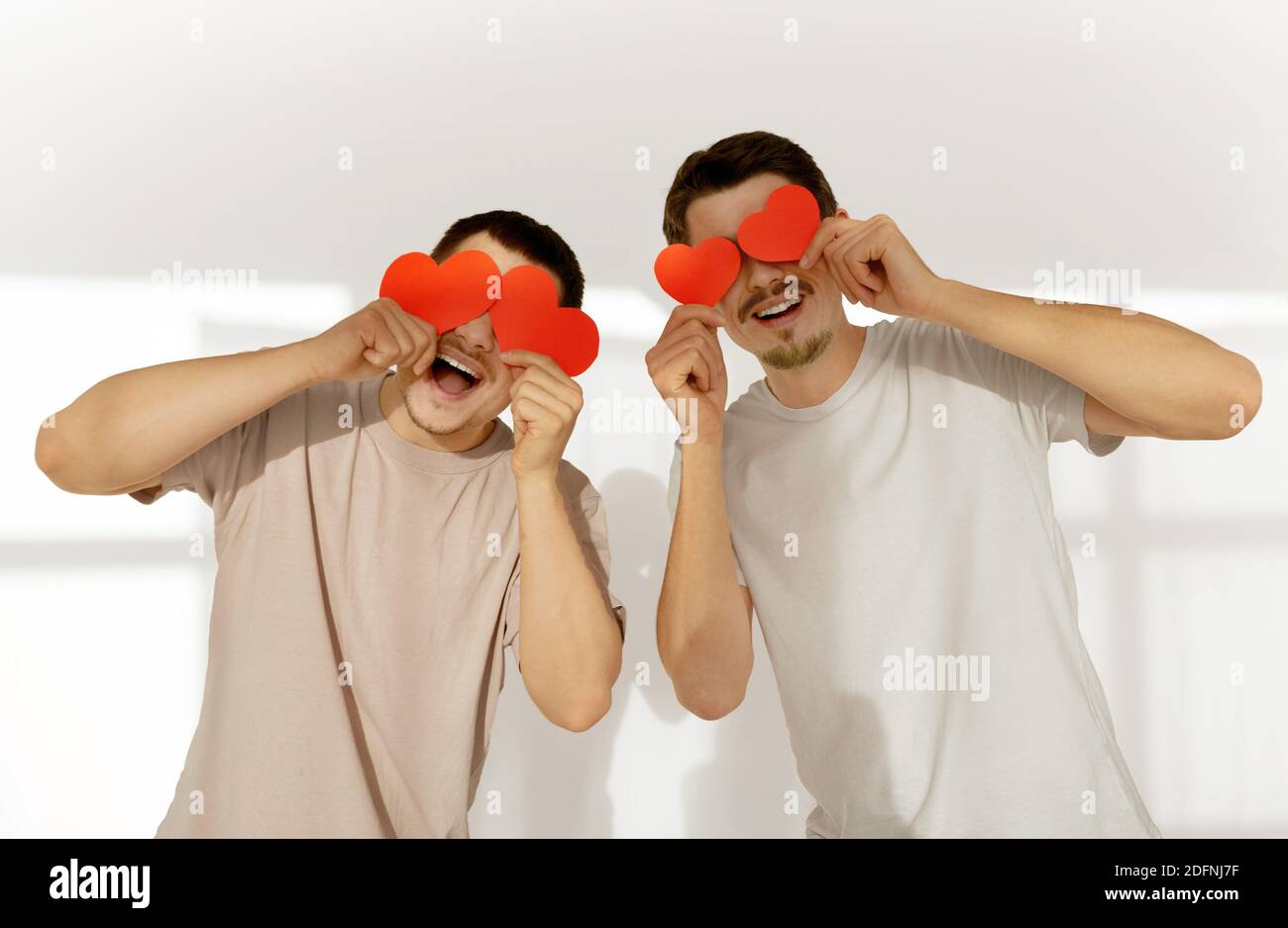 Happy couple of young gay men hide their eyes behind red paper valentine hearts in celebration at home against white wall background Stock Photo