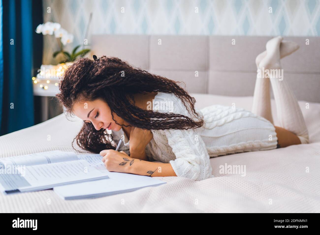 Young afro hair mix raced woman in home clothes lie on bed and has an online lesson with tablet. Distance learning or work concept, Stock Photo