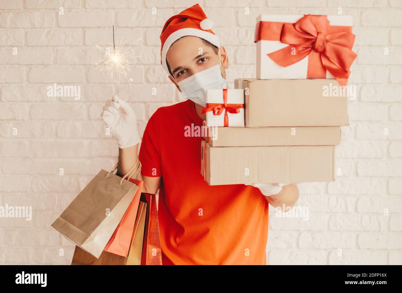 https://c8.alamy.com/comp/2DFP16X/young-festive-delivery-man-in-medical-face-mask-protective-gloves-santa-hat-hold-light-sparkler-shopping-bags-cardboard-gift-boxes-and-smile-happ-2DFP16X.jpg