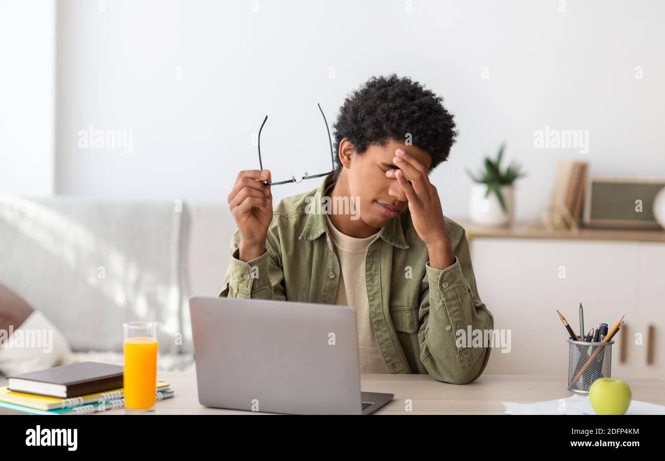 Black teenager feeling tired, rubbing irritated eyes, sitting at desk with laptop, exhausted from studying online Stock Photo