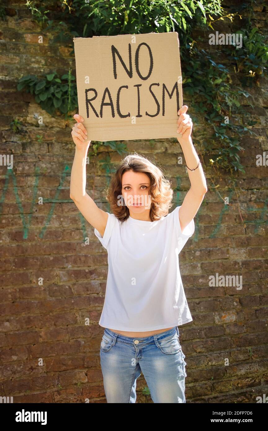 Upset young protesting woman in white shirt holds protest sign broadsheet placard with slogan 'No racism' for public demonstration on stone wall Stock Photo