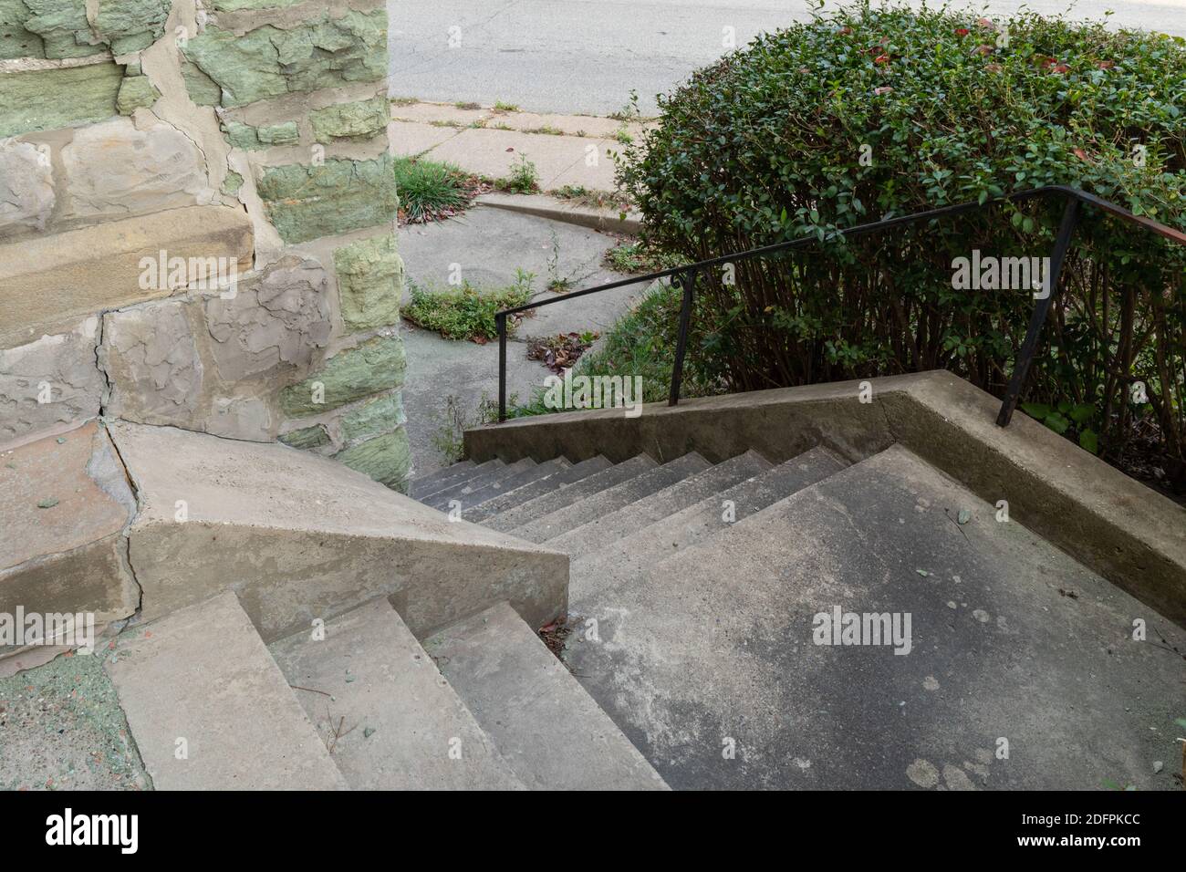 Concrete stairs descending around an outside corner of a building made from crumbling green limestone, black handrails on steep descent, horizontal as Stock Photo