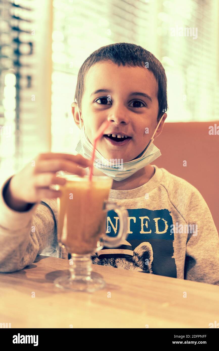 A child wearing a mask in a cafe during a pandemic. A boy in a cafe drinks sea buckthorn tea in a cafe during a pandemic. toned Stock Photo