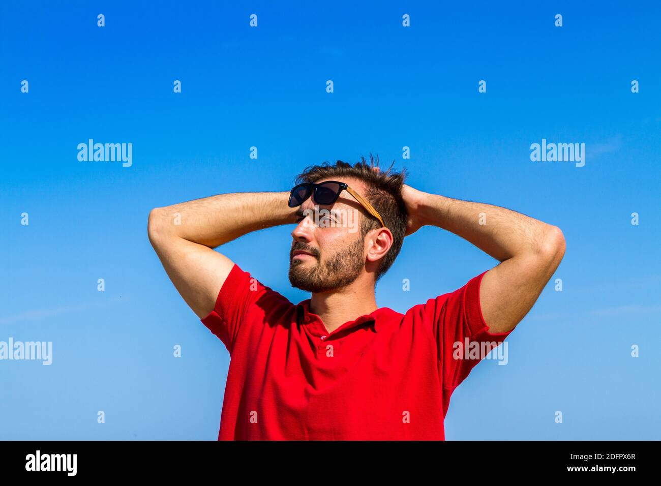 Young man expressive dynamic ambitious successful bearded in red t-shirt sunglasses in summer blue sky background. Stock Photo
