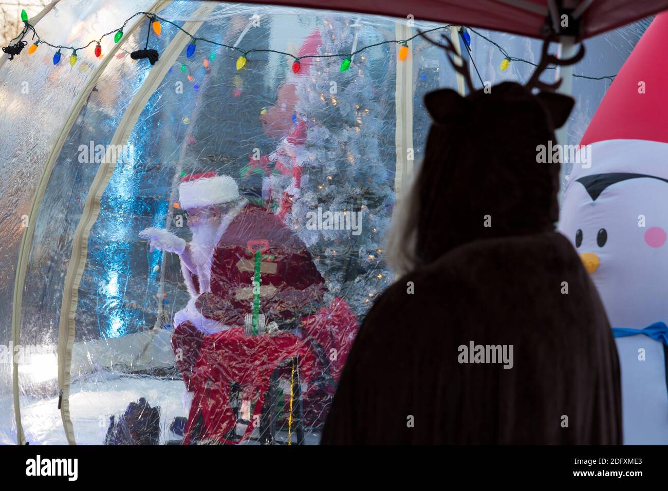 Seattle, Washington, USA. 6th December, 2020. Assistant LaRae Weidner looks on as Dan Kemmis, known as the Seattle Santa, greets visitors from within a protective plastic bubble in Seattle’s Greenwood neighborhood. Unable to schedule private events due to the coronavirus pandemic, Kemmis is making physically distanced public appearances with some donations benefiting local homeless shelters. Credit: Paul Christian Gordon/Alamy Live News Stock Photo