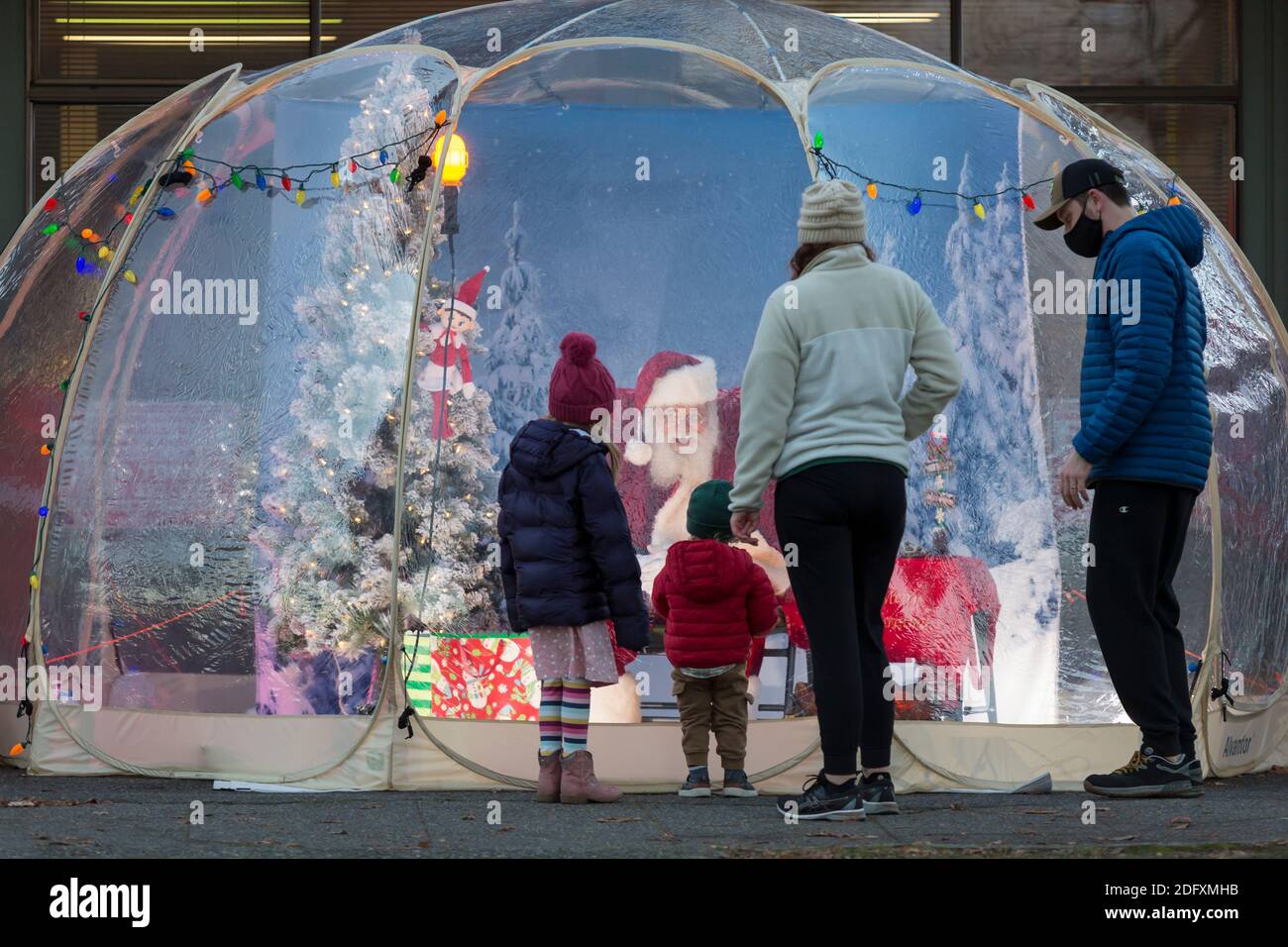 Seattle, Washington, USA. 6th December, 2020. Dan Kemmis, known as the Seattle Santa, greets a young family from within a protective plastic bubble in Seattle’s Greenwood neighborhood. Unable to schedule private events due to the coronavirus pandemic, Kemmis is making physically distanced public appearances with some donations benefiting local homeless shelters. Credit: Paul Christian Gordon/Alamy Live News Stock Photo