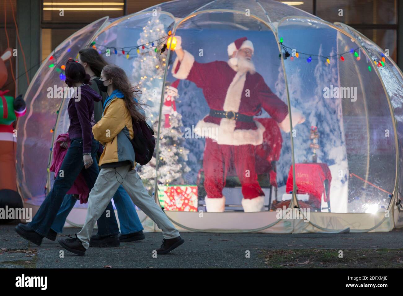 Seattle, Washington, USA. 6th December, 2020. Dan Kemmis, known as the Seattle Santa, stretches as a group of young women walk by his protective plastic bubble in Seattle’s Greenwood neighborhood. Unable to schedule private events due to the coronavirus pandemic, Kemmis is making physically distanced public appearances with some donations benefiting local homeless shelters. Credit: Paul Christian Gordon/Alamy Live News Stock Photo
