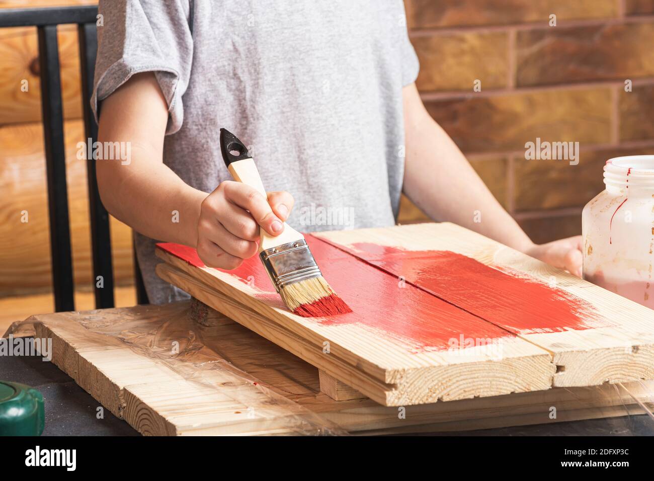 A boy paints a wooden fake with a paintbrush in red in a country house. DIY painting the walls. Boy artist doing home renovations. Stock Photo