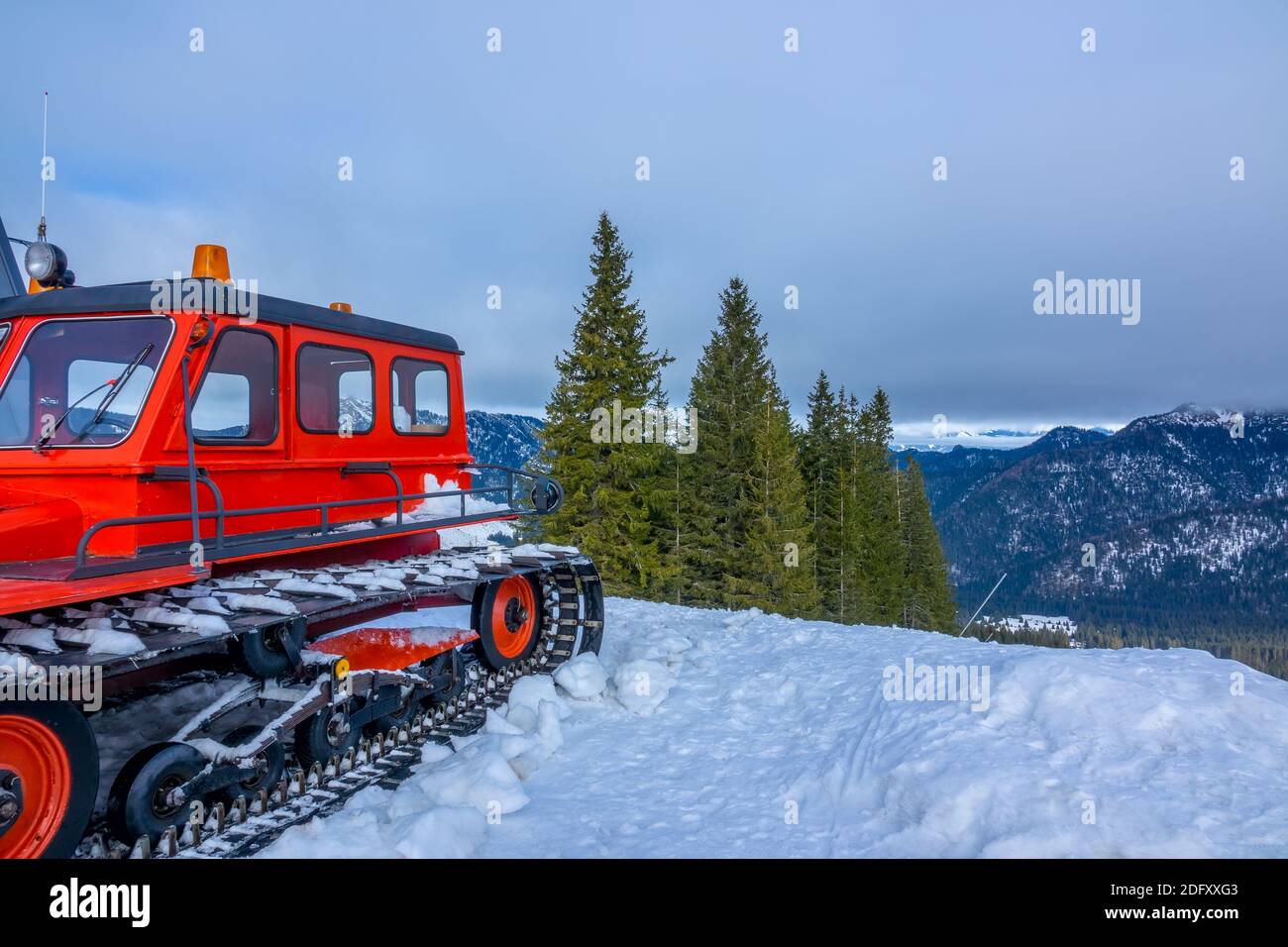 Cloudy sky over snow-capped mountain peaks. Wooded slopes. Red snowcat in the foreground Stock Photo