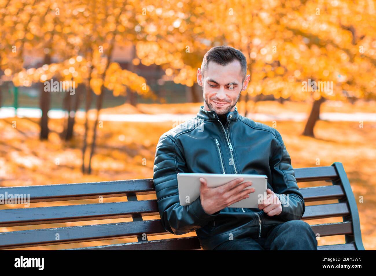 Handsome Young Man Sitting on the Bench and Using Big White Tablet PC at the Beautiful Autumn Park Stock Photo