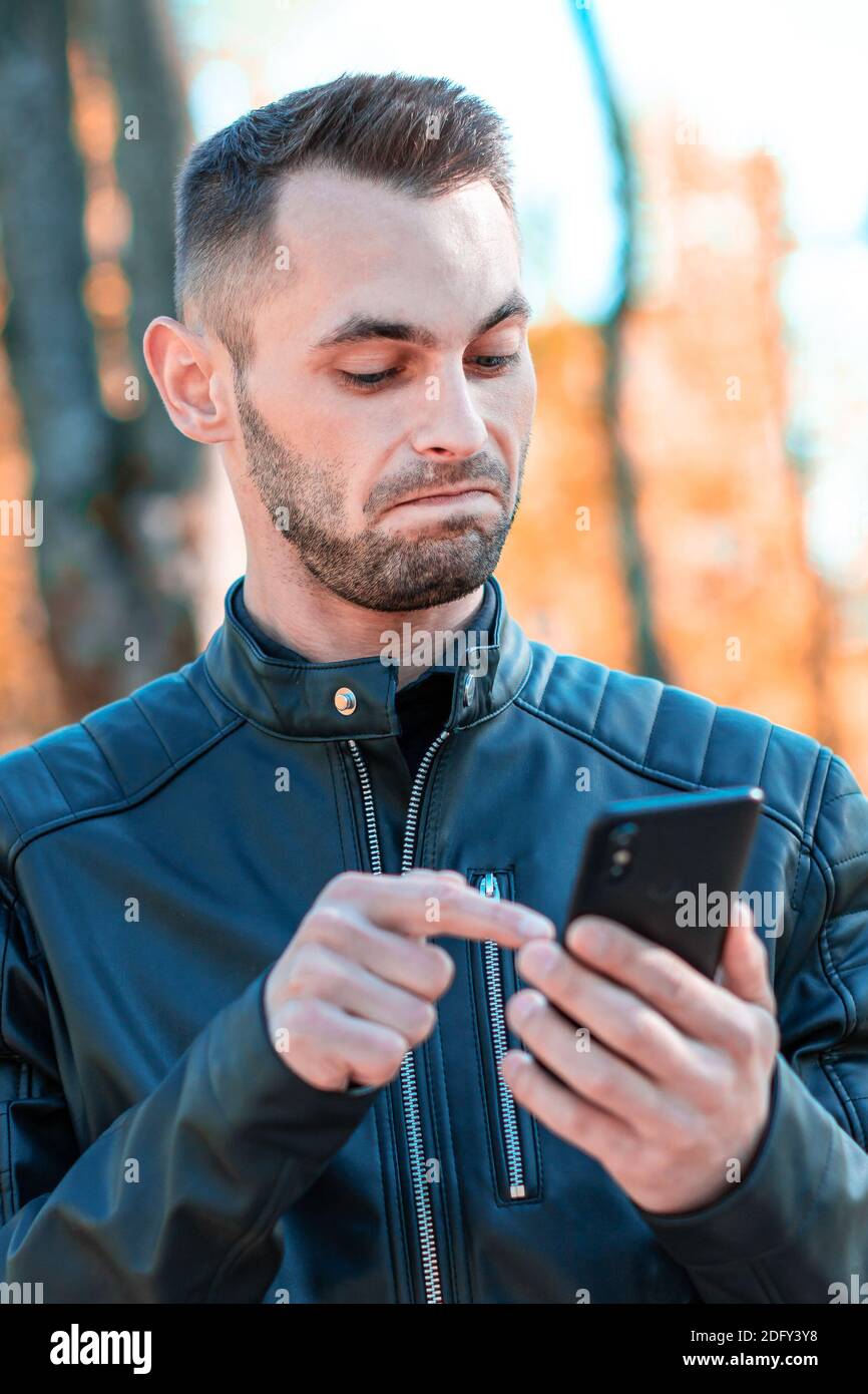 Youthful Guy Was Surprised Using Black Smartphone at the Beautiful Autumn Park. Handsome Young Man with Mobile Phone at Sunny Day - Medium CloseUp Portrait Stock Photo