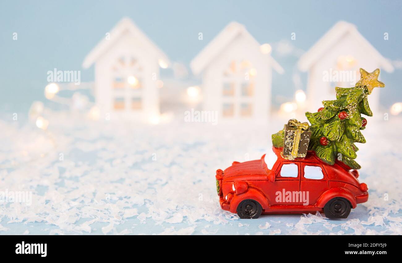 Christmas decor - red retro car on snow carries past white houses with lights garlands in bokeh Christmas tree with gift boxes on roof. Toy on blue ba Stock Photo