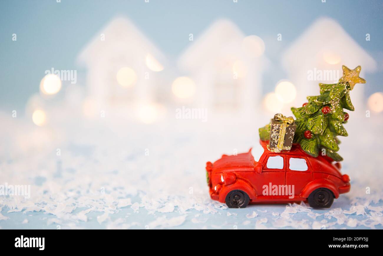 Christmas decor - red retro car on snow carries past white houses with lights garlands in bokeh Christmas tree with gift boxes on roof. Toy on blue ba Stock Photo