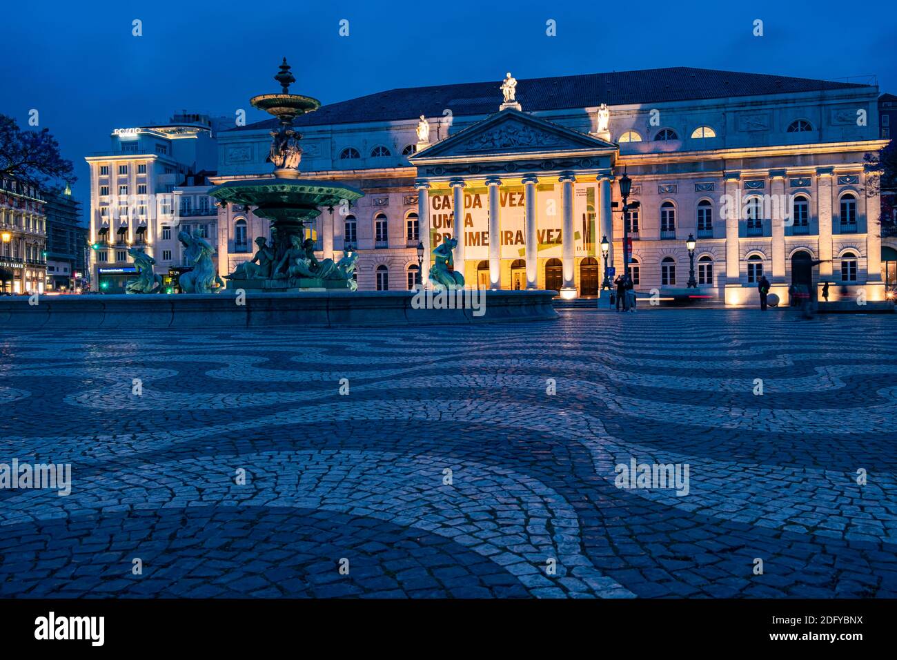 Low angle shot of fountain and illuminated facade of National Theatre as people walking at Rossio Square in Lisbon, Portugal Stock Photo