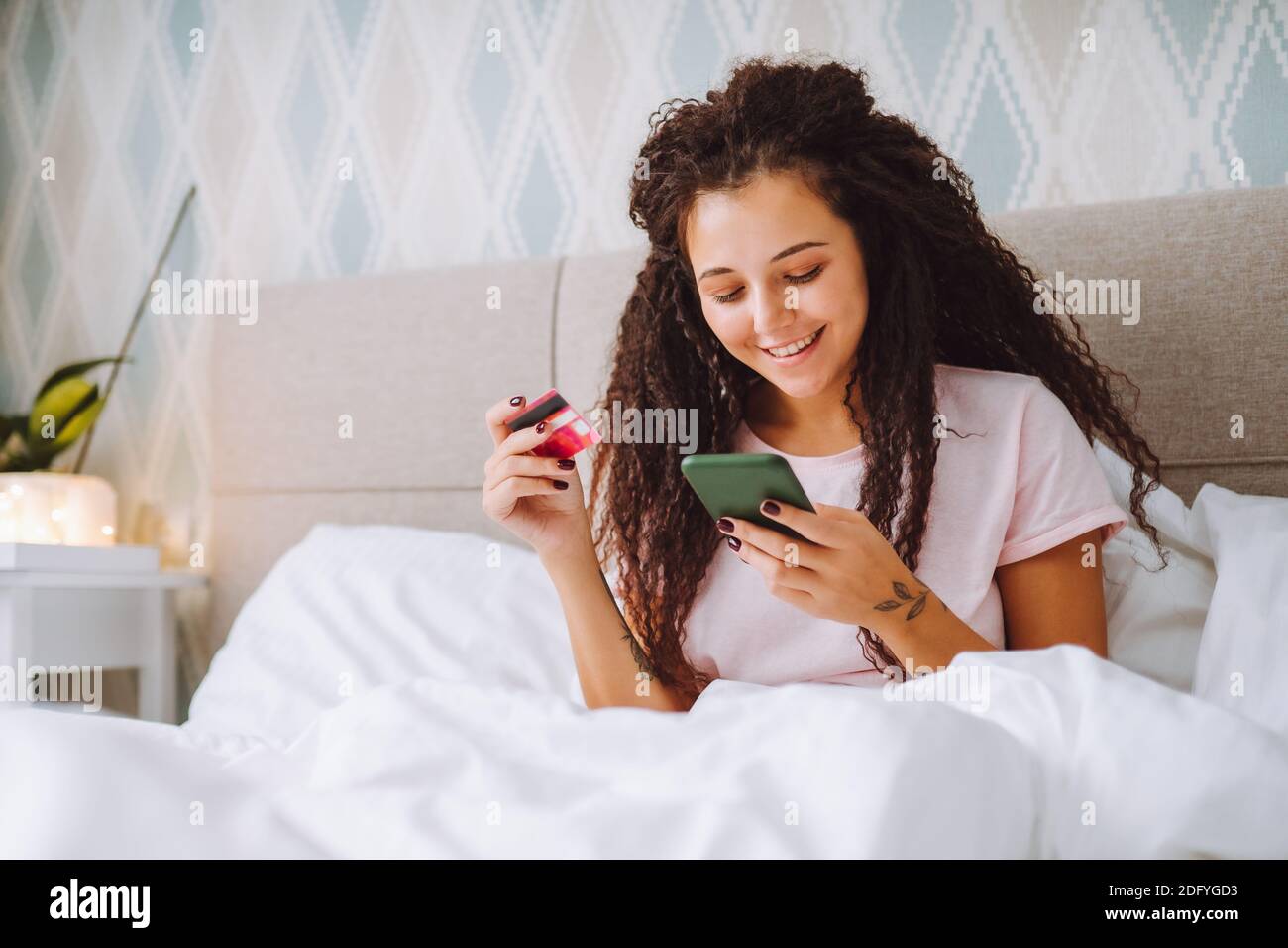 Positive young afro haired woman shop online using her smartphone sitting at home in bed paying with bank card Stock Photo