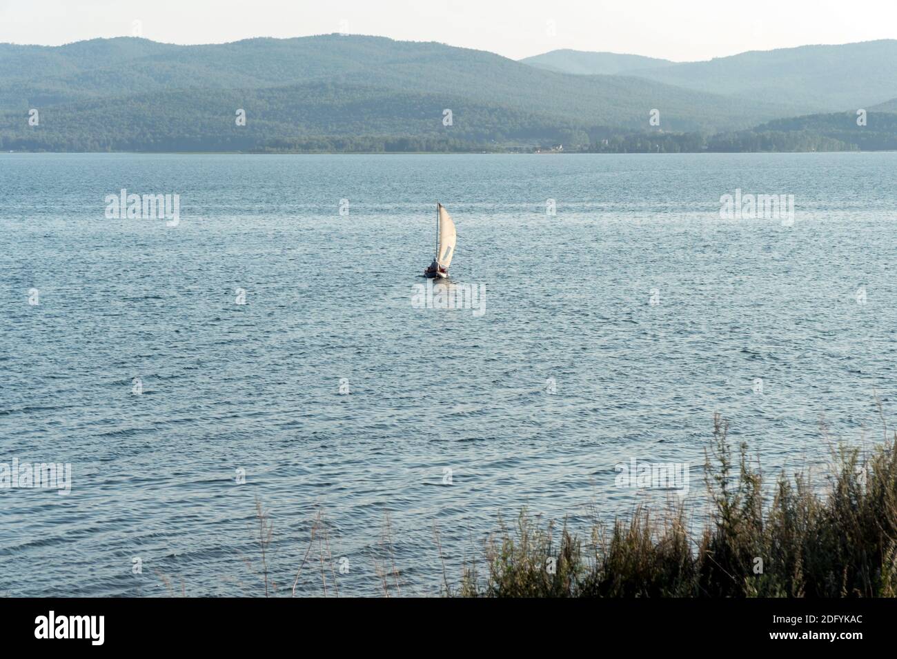 An inflatable boat with a sail floats on the water surface of the lake against the backdrop of wooded mountains on a sunny summer day. Stock Photo