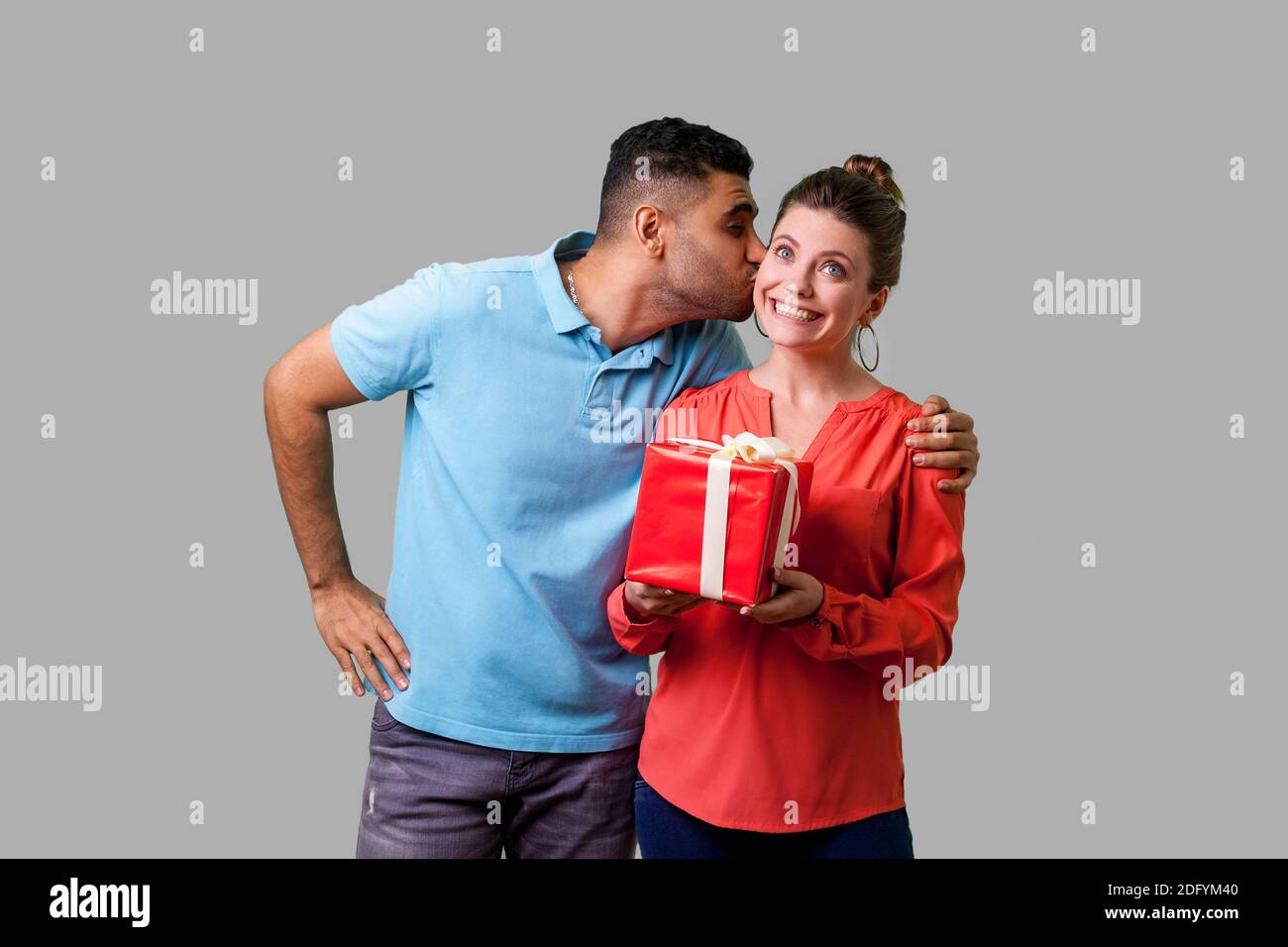 Family celebrating Valentines day. Young man in casual clothes kissing woman on cheek, she's holding gift box and looking up dreamy and surprised. iso Stock Photo