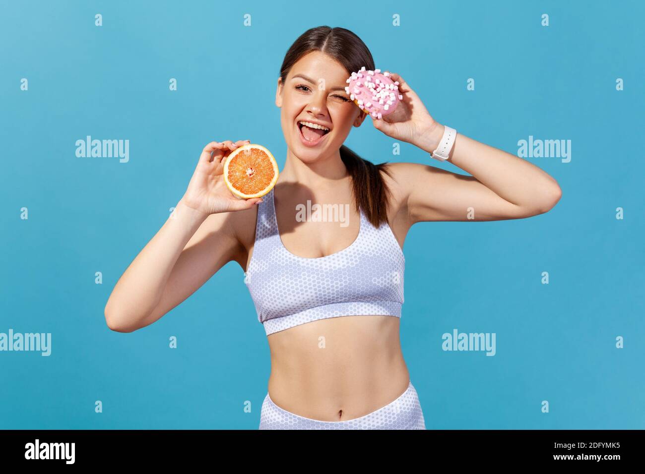 Positive athletic woman in white sportswear holding half of ripe juicy grapefruit and round donut with pink icing, having fun looking camera with smil Stock Photo