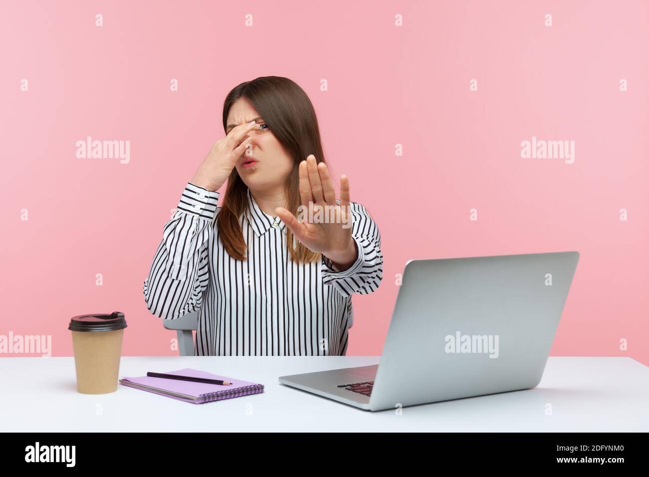 Confused woman office employee sitting at workplace with laptop and holding breath, pinching her nose to avoid awful stink, gesturing stop. Indoor stu Stock Photo