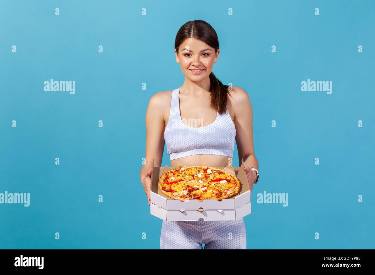 Portrait of positive sports woman holding cardboard box with fatty pizza, enjoying junk food after workout in gym, lack of willpower. Indoor studio sh Stock Photo
