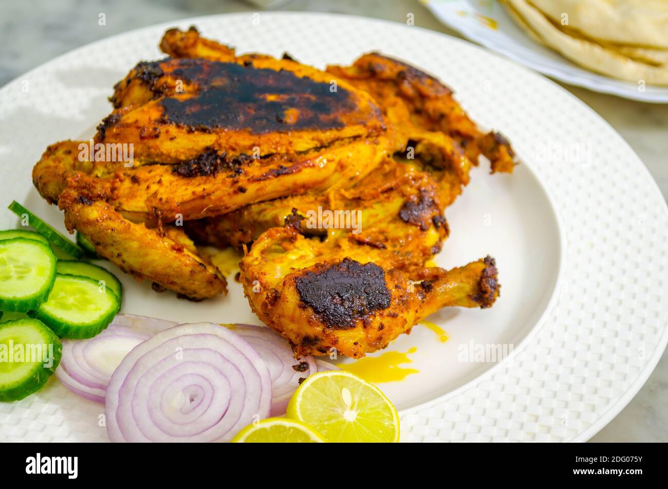 Beautifully placed ready-to-eat full chicken tandoori on a white plate along with sliced onions, cucumbers and lemon Stock Photo