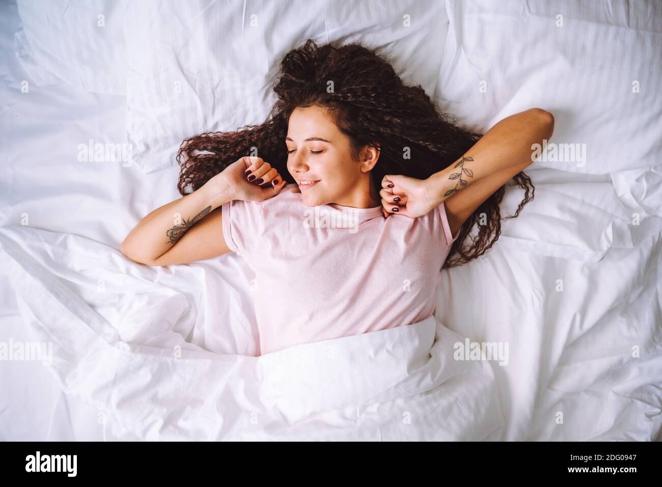 Top view of young woman waking up in early morning lying on bed with snow white linens and sretch her arms and body with smile Stock Photo