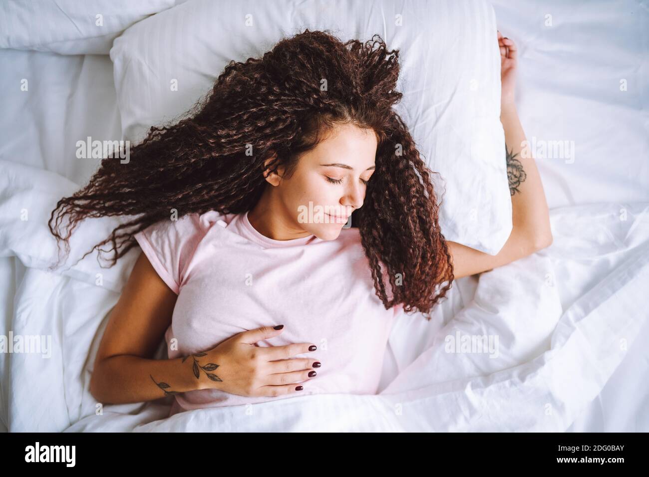 Top view of young afro haired woman sleeping in morning lying on bed with snow white linens. Stock Photo
