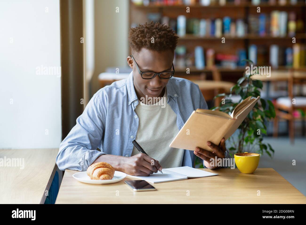 Funky black guy with textbook taking notes in copybook at cozy cafe ...