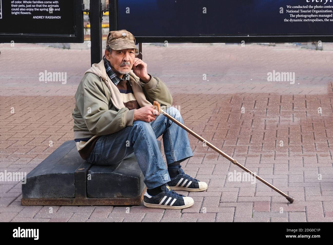 Frustrated Sad Elderly Man with stick sitting on street in solitude and looking at camera. Poverty and Despair, Social Pension Problems Stock Photo
