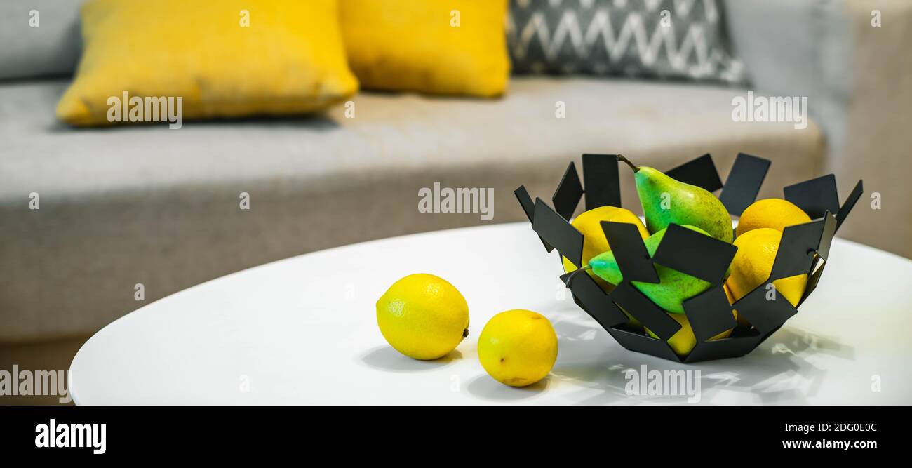 Close-up of decorative bowl with fruits on white table. Lemons and pears. Modern interior. Cozy sofa with cushions. Stock Photo
