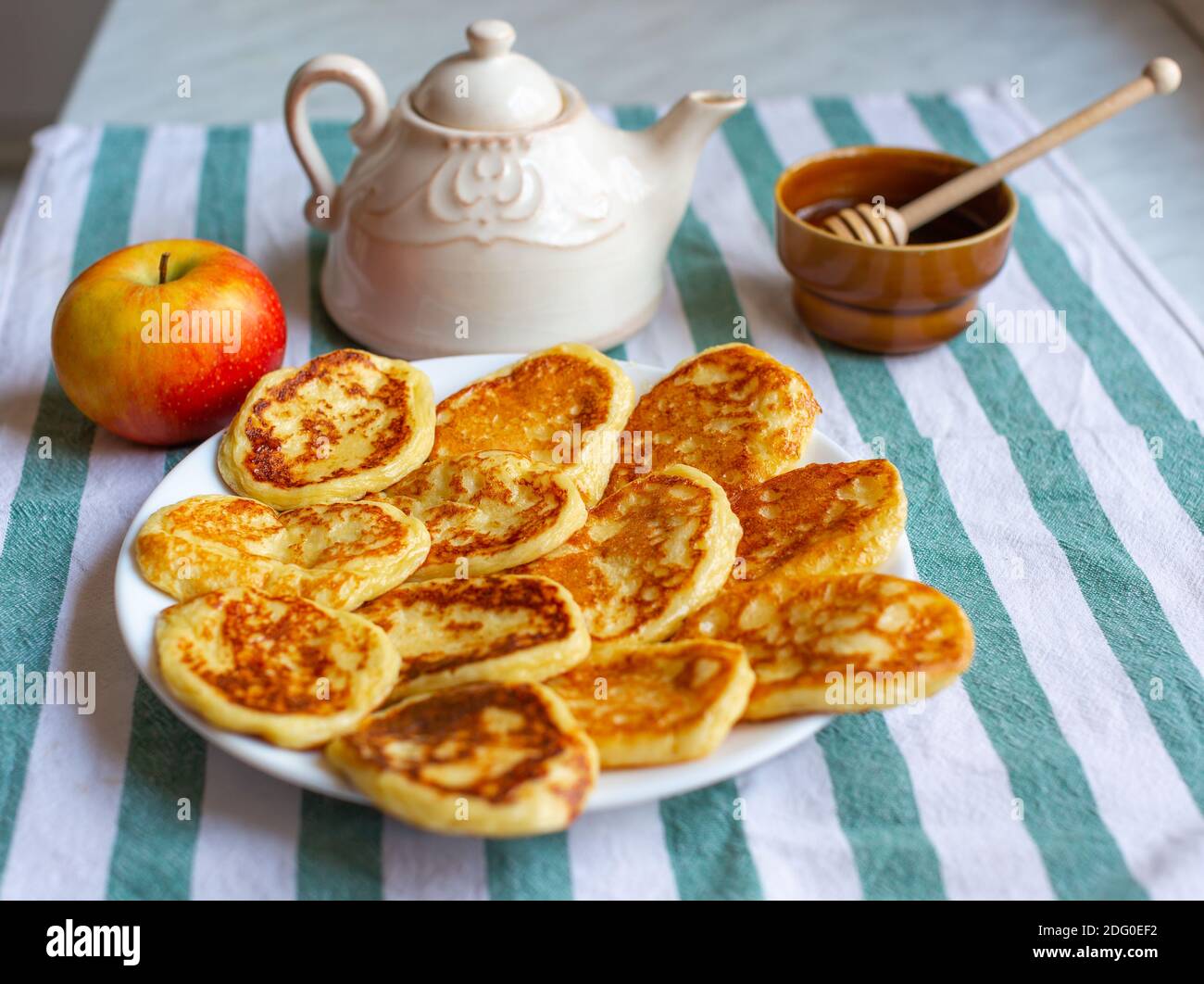 Tasty homemade pancakes laid out on plate near an apple, teapot and bowl of honey . Selective focus on pancakes Stock Photo