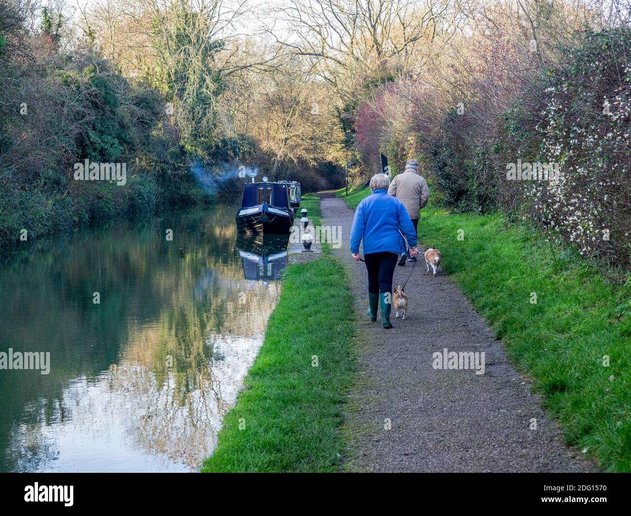 Winter scene on the Grand Union canal, Stoke Bruerne, Northamptonshire, UK; two dog walkers passing a narrowboat with smoking chimney Stock Photo