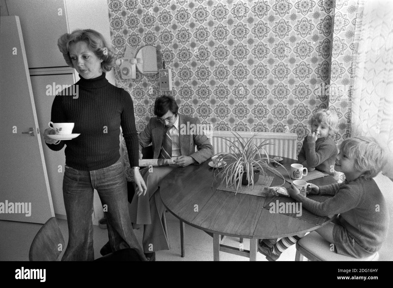 1970s England, a middle income professional couple at home with their two children. In the kitchen, mother making afternoon tea, sandwiches for the two boys, and a cup of tea for her husband who is reading the newspaper and paying no attention at all or interest in his children. He is bored family life. 1977 New town modern housing development Milton Keynes Buckinghamshire UK HOMER SYKES Stock Photo